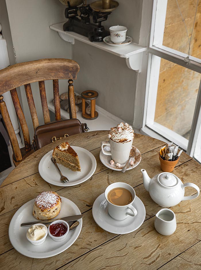 A cozy table at Lucy's Tearoom in Stow-on-the-Wold, Cotswolds, is set with a slice of cake, a scone with clotted cream and jam, a cup of tea, and a mug of hot chocolate topped with whipped cream and marshmallows. The rustic wooden table and vintage decor add to the charming, quaint atmosphere.