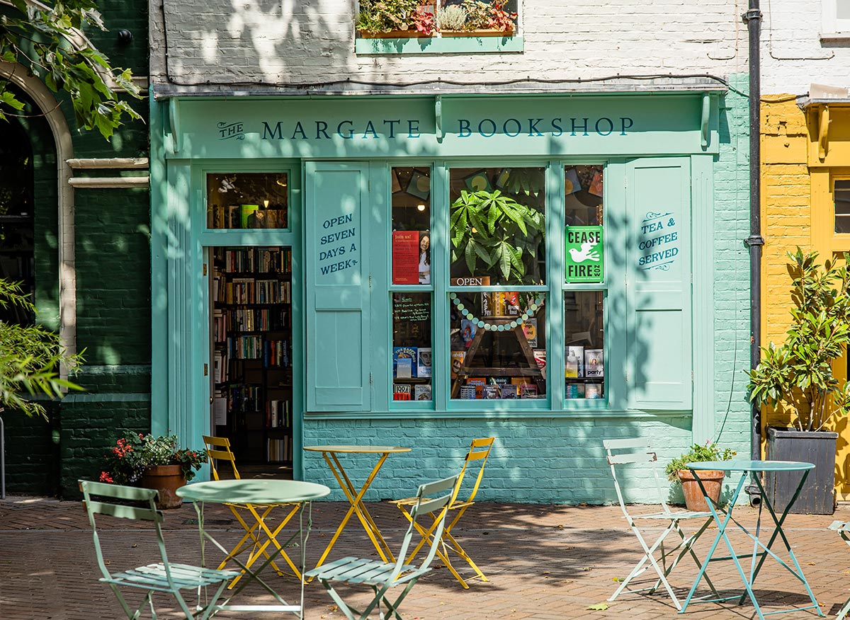 The storefront of The Margate Bookshop, featuring a light blue facade with large windows displaying books and plants. The sign reads "Open Seven Days a Week" and "Tea & Coffee Served". Outside, there are colorful yellow and blue chairs and tables, creating a welcoming atmosphere for visitors to sit and enjoy a book or a drink. The scene is set on a sunny day with dappled light from nearby trees.