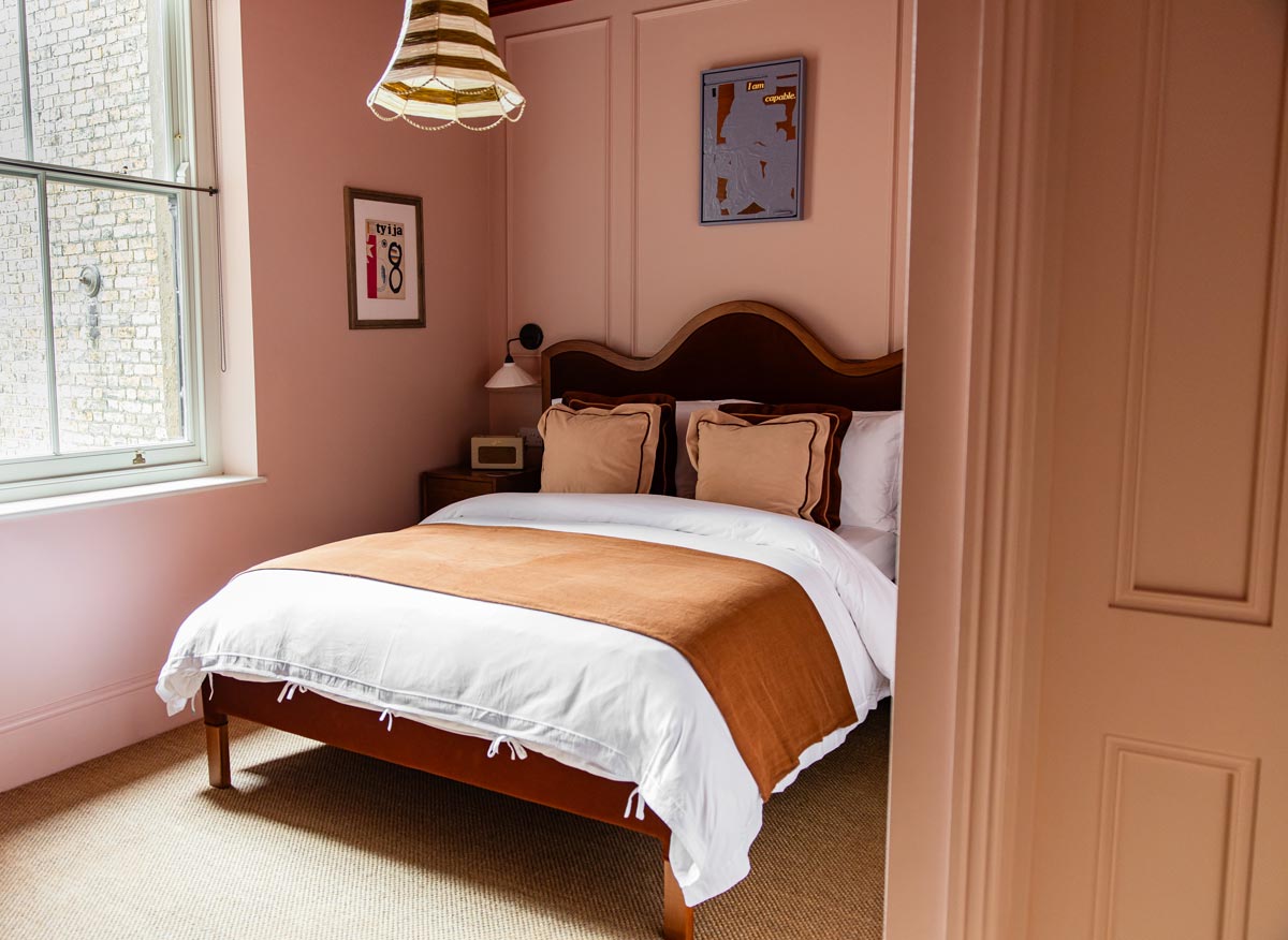 A cozy bedroom with soft pink walls and a wooden bed frame topped with white and brown bedding. The room features a large window, a small nightstand with a lamp, and framed artwork on the walls. A striped pendant light hangs from the ceiling, adding a touch of charm to the space.






