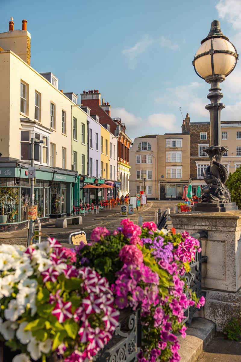 A charming street scene in Margate's Old Town, showcasing a row of colorful buildings housing cafes and shops, including The Margate Coffee Shed. The street is adorned with vibrant flowers in the foreground, and classic street lamps add to the picturesque setting. Outdoor seating and clear skies create a welcoming and lively atmosphere.