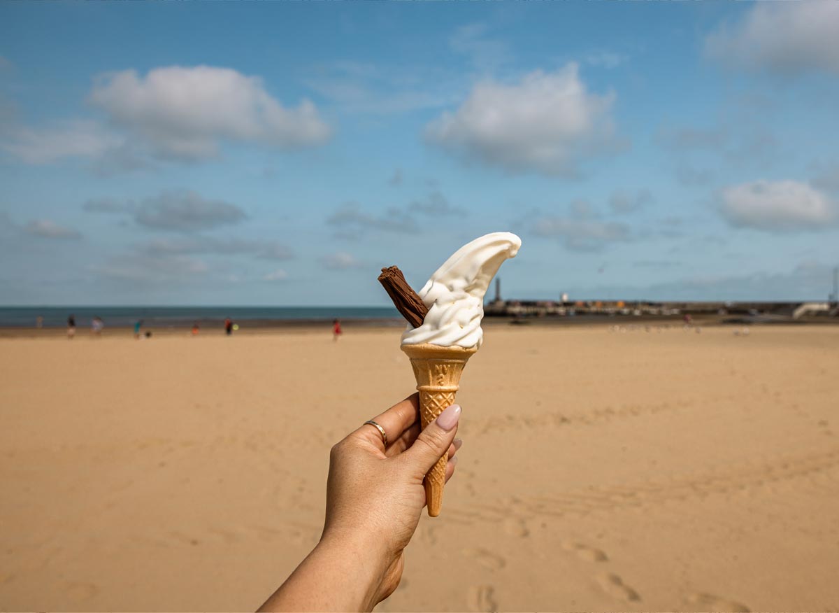 A hand holding a soft serve ice cream cone with a chocolate flake, set against the backdrop of a sandy beach and the ocean. The sky is partly cloudy, and there are a few people in the distance enjoying the beach. The scene captures a quintessential seaside moment, evoking a sense of summer relaxation.