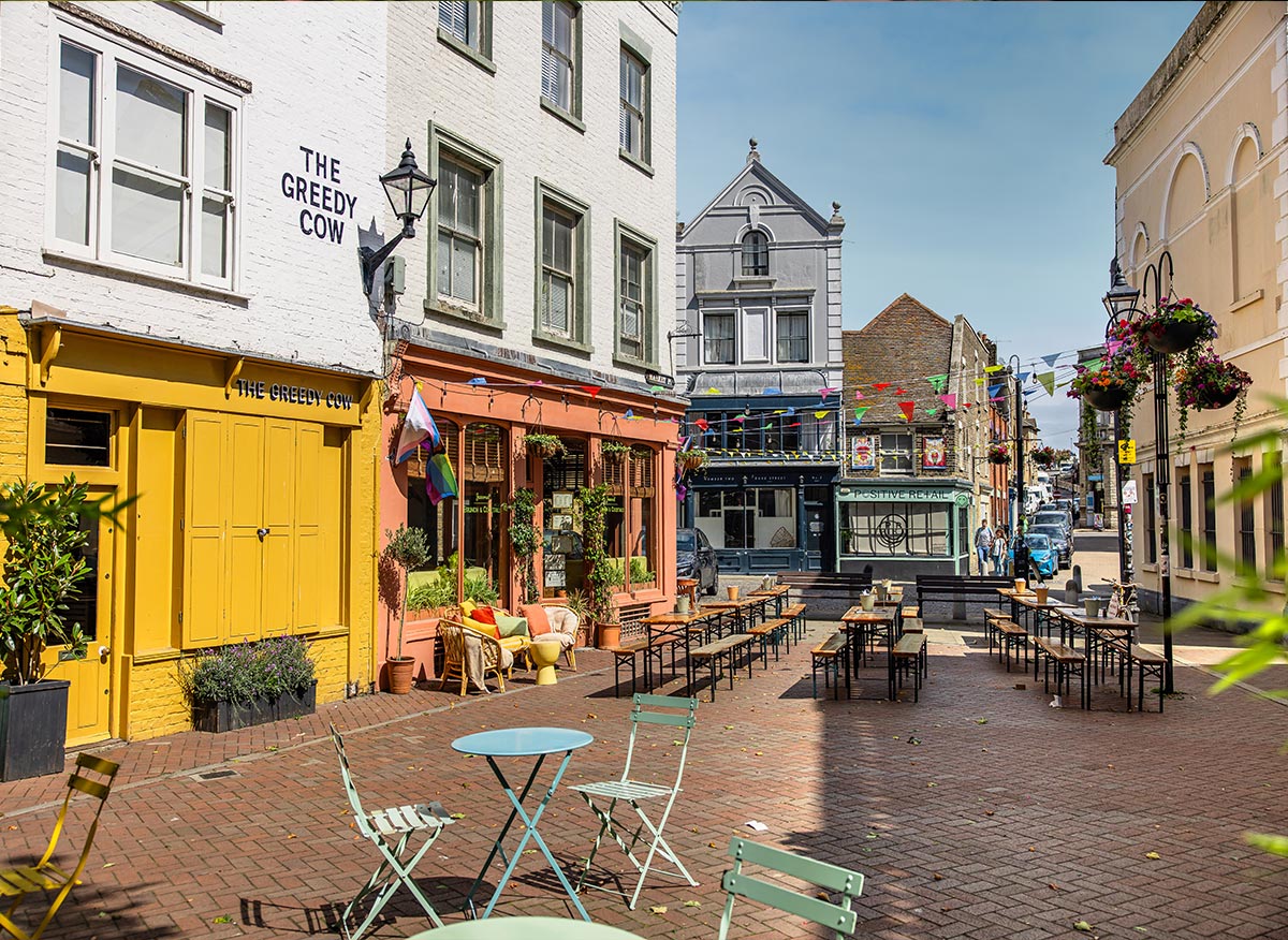 A lively scene in Margate's Old Town Square, featuring colorful buildings and outdoor seating. The Greedy Cow, with its bright yellow facade, and adjacent cafes create a welcoming atmosphere. The square is decorated with festive bunting and filled with tables and chairs for diners, with vibrant plants and flowers adding to the cheerful ambiance. The picturesque setting invites visitors to relax and enjoy the charming surroundings.






