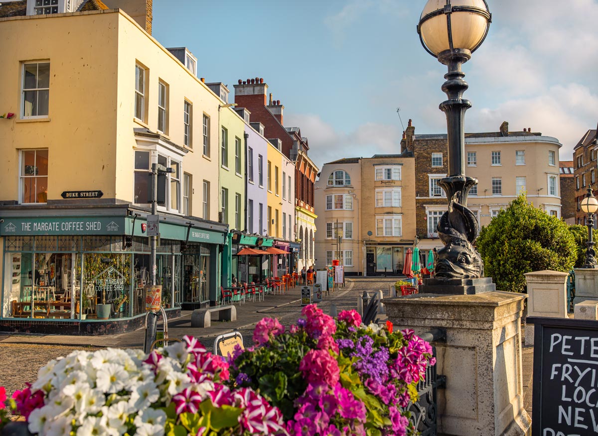 A charming street scene in Margate's Old Town, showcasing a row of colorful buildings housing cafes and shops, including The Margate Coffee Shed. The street is adorned with vibrant flowers in the foreground, and classic street lamps add to the picturesque setting. Outdoor seating and clear skies create a welcoming and lively atmosphere.