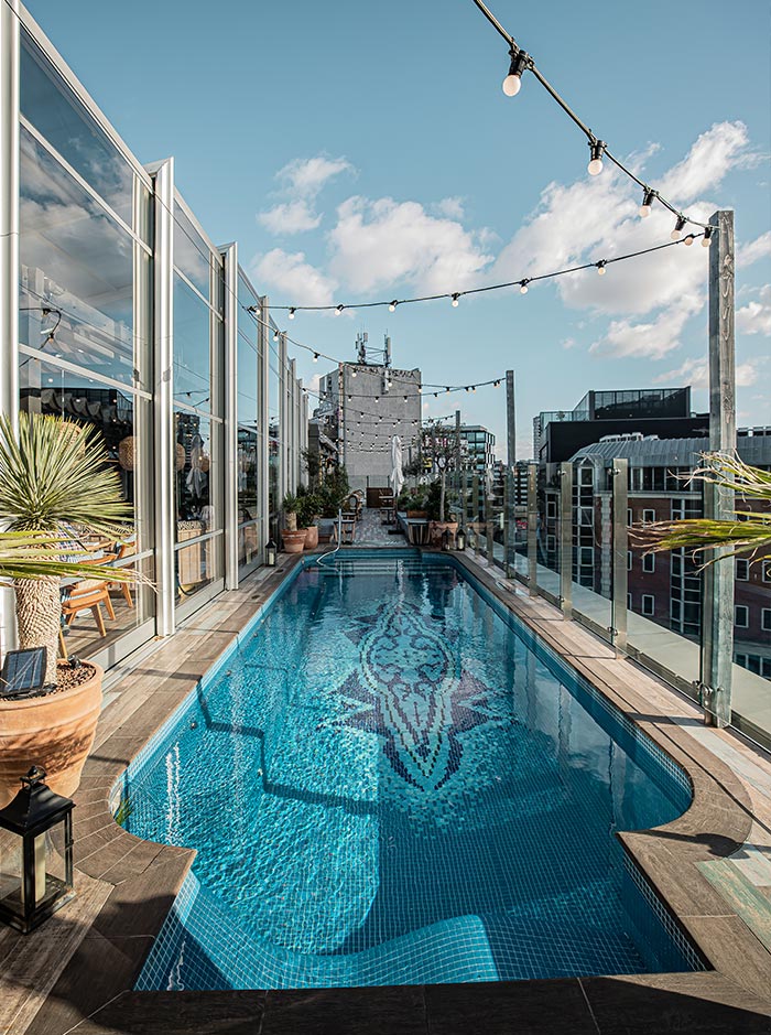 A chic rooftop pool at the Mondrian Hotel in London, featuring a long, narrow pool with a decorative mosaic design at the bottom. The pool area is lined with glass railings, potted plants, and outdoor seating, offering stunning views of the city skyline. String lights hang above, adding a touch of ambiance against the blue sky and scattered clouds.