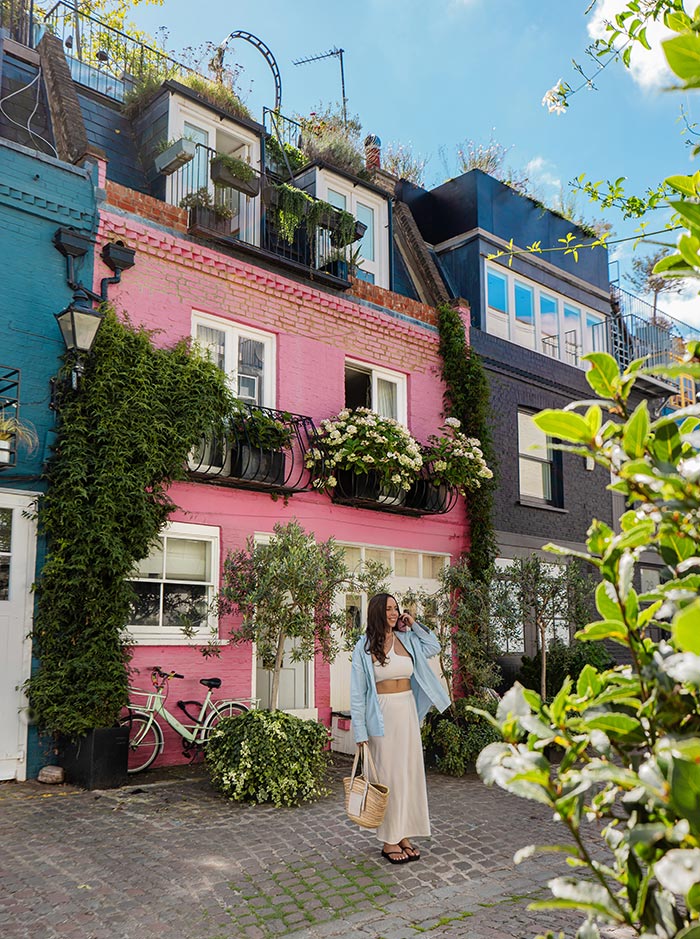 A woman in a beige dress and light blue jacket stands in front of a vibrant pink house in Notting Hill, London. The house is adorned with lush greenery and blooming flowers on the balconies, creating a picturesque and charming scene reminiscent of the "Love Actually" movie. A green bicycle and potted plants add to the quaint, cozy atmosphere.