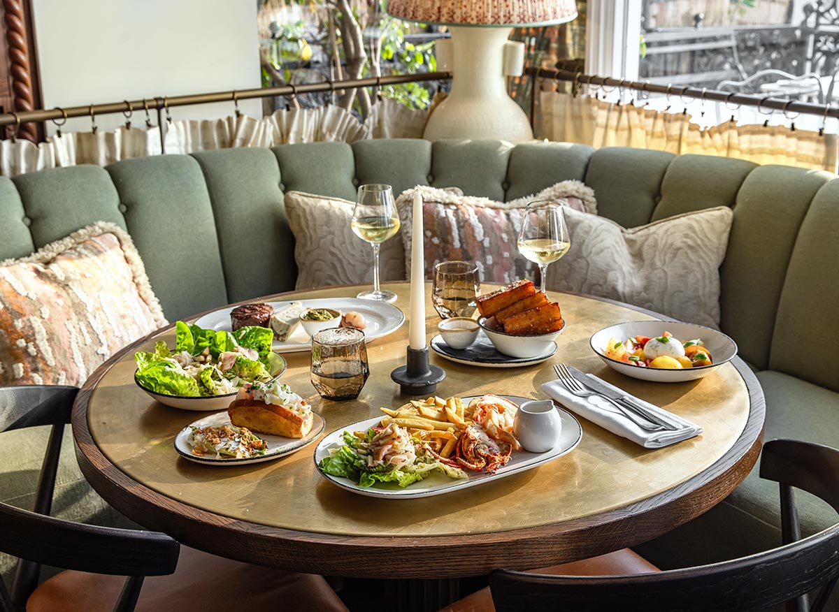 A cozy dining setup at Pearly Cow in No. 42 Margate, featuring a round table with an assortment of delicious dishes. The table is set with plates of salad, fries, seafood, and other gourmet items, accompanied by glasses of white wine. The setting includes a comfortable green sofa with decorative pillows and a warm, inviting atmosphere enhanced by natural light streaming in from the window.