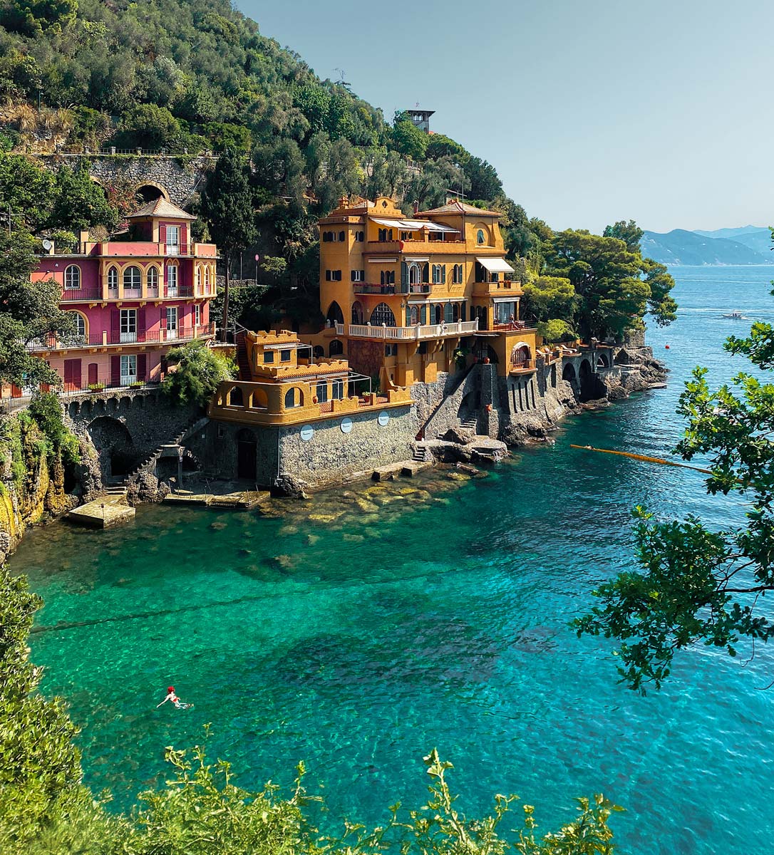 A stunning view of Baia del Cannone in Portofino, Italy, showcasing colorful buildings perched on the cliffs above clear turquoise waters. A lone swimmer enjoys the serene sea, surrounded by lush greenery and scenic hills.






