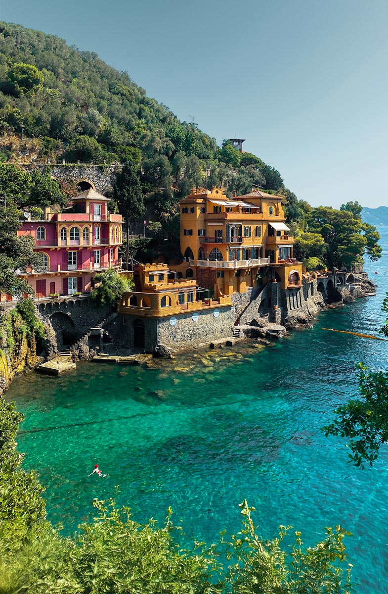 A stunning view of Baia del Cannone in Portofino, Italy, showcasing colorful buildings perched on the cliffs above clear turquoise waters. A lone swimmer enjoys the serene sea, surrounded by lush greenery and scenic hills.