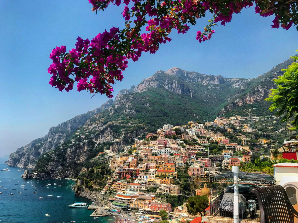 A stunning view of Positano, Italy, with vibrant pink bougainvillea flowers framing the image. The hillside is adorned with colorful buildings cascading down towards the azure sea, where numerous boats are anchored near the shore. The lush green mountains provide a dramatic backdrop to this picturesque coastal town.






