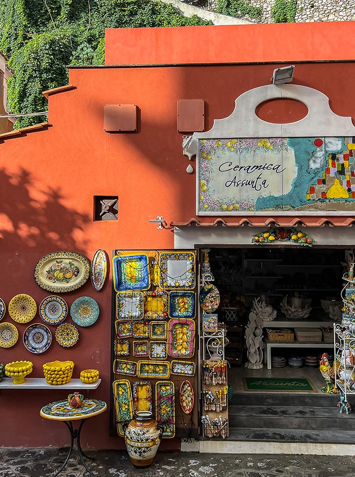 A vibrant ceramic shop in Positano, Italy, with a bold orange exterior displaying an array of colorful hand-painted plates, trays, and other pottery items. The shop sign reads "Ceramica Assunta," and the entrance showcases more intricate ceramic pieces and decorative items, inviting visitors to explore the artisanal crafts inside.