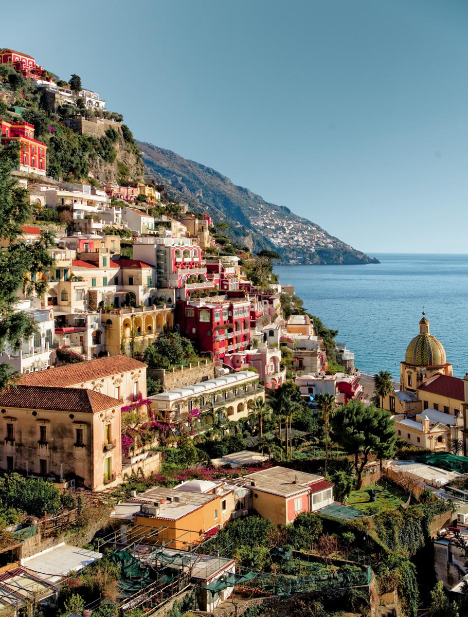 A scenic view of Positano, Italy, showcasing the colorful buildings stacked along the steep hillside, leading down to the tranquil blue waters of the Mediterranean Sea. Prominent in the scene is the dome of the Church of Santa Maria Assunta, surrounded by lush greenery and vibrant bougainvillea. The clear sky enhances the beauty of this charming coastal village.