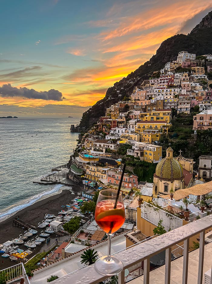 A picturesque view of Positano, Italy at sunset, featuring colorful hillside buildings and a serene coastline with boats and beach umbrellas. In the foreground, a glass of Aperol Spritz sits on a balcony railing, adding a touch of relaxation to the scenic vista.