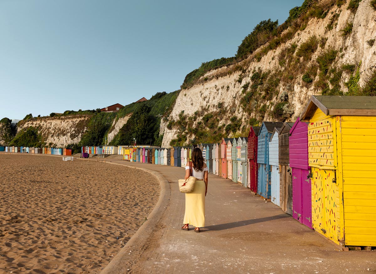 A woman wearing a white top and a yellow skirt walks along a pathway by the beach, lined with colorful beach huts. The huts are painted in various vibrant colors and are situated at the base of a steep, grassy cliff. The sandy beach extends to the left, with a clear blue sky overhead.