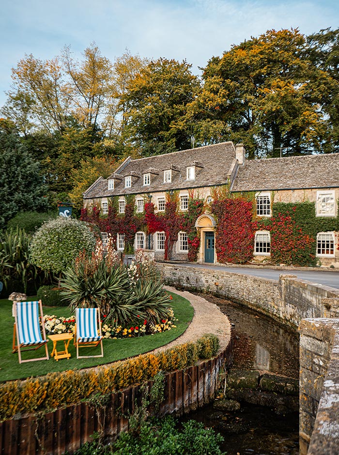 The Swan Inn in the Cotswolds, a charming stone building covered in vibrant ivy, sits alongside a serene stream. Two striped deck chairs and a small yellow table are set up on a manicured lawn, inviting relaxation in the picturesque setting. The surrounding trees show hints of autumn, enhancing the inn's cozy and inviting atmosphere.