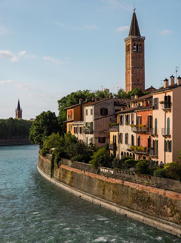 A picturesque view of the Adige River in Verona, lined with colorful buildings adorned with balconies and lush greenery. The scene is dominated by a tall bell tower rising above the rooftops, with another tower visible in the distance. The river winds gracefully through the city, bathed in the soft, golden light of late afternoon, creating a serene and charming atmosphere.