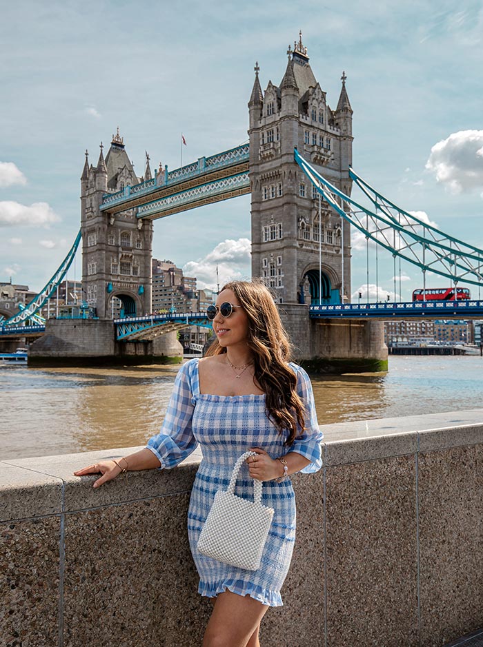 A woman in a blue and white checkered dress stands beside the Thames River, with Tower Bridge prominently in the background. The iconic bridge, featuring its two towers and blue suspension elements, creates a picturesque backdrop against a partly cloudy sky. The woman, accessorized with sunglasses and a pearl handbag, adds a fashionable touch to the scenic London landmark.