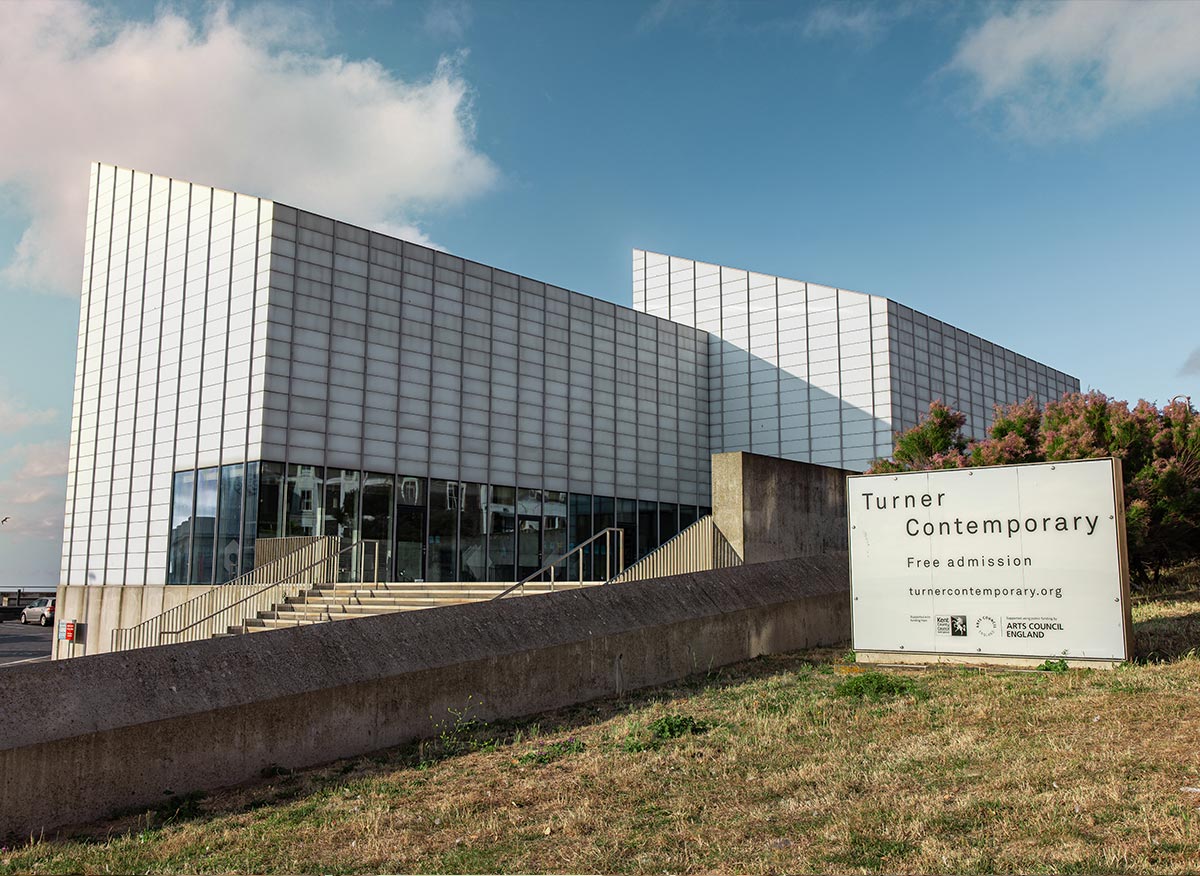 The modern, sleek exterior of the Turner Contemporary art gallery in Margate, featuring its distinctive geometric architecture with a glass facade. A sign near the entrance reads "Turner Contemporary - Free admission" along with the website and Arts Council England logo. The building is set against a backdrop of a clear blue sky with a few clouds, emphasizing its contemporary design and inviting atmosphere.