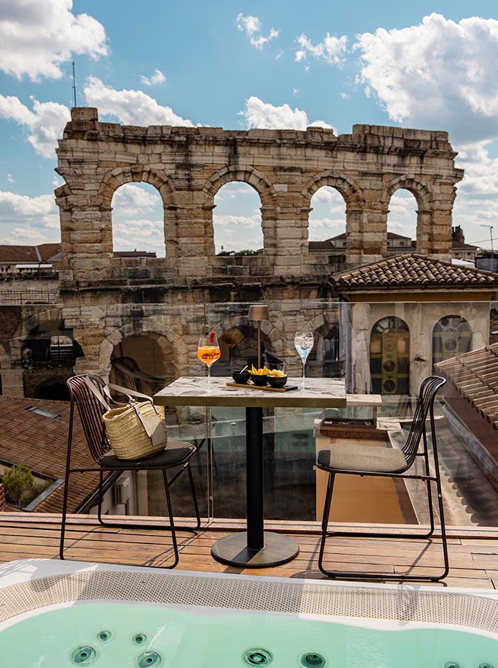 A stylish rooftop terrace in Verona, featuring a table set with drinks and snacks, overlooks the ancient Roman amphitheater, Arena di Verona. The scene includes two chairs, one with a straw bag, and a hot tub in the foreground. The historic stone arches of the amphitheater provide a striking contrast to the modern, comfortable setting, under a bright, partly cloudy sky.