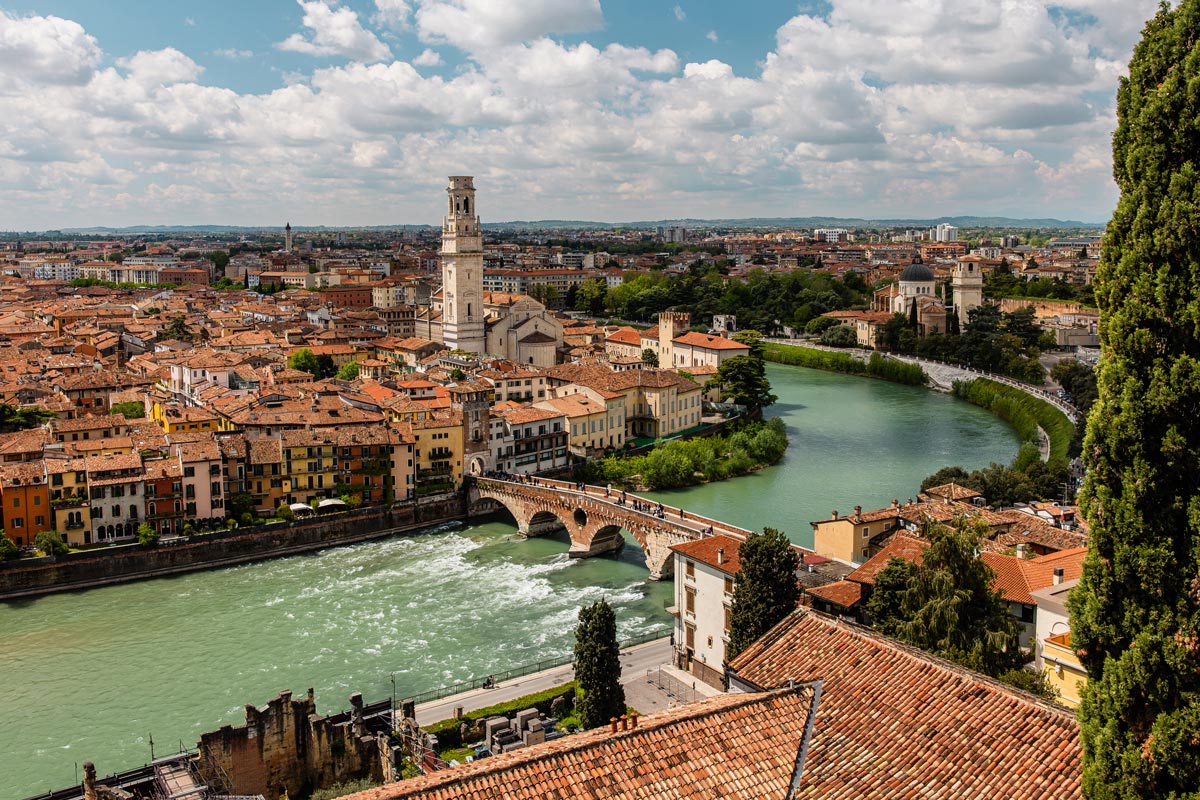A panoramic view of Verona, showcasing the Adige River winding through the historic city. The scene includes the iconic Ponte Pietra bridge, surrounded by charming buildings with terracotta roofs, and the prominent Verona Cathedral with its tall bell tower. The picturesque landscape is bathed in sunlight under a partly cloudy sky, highlighting the blend of architectural beauty and natural scenery.