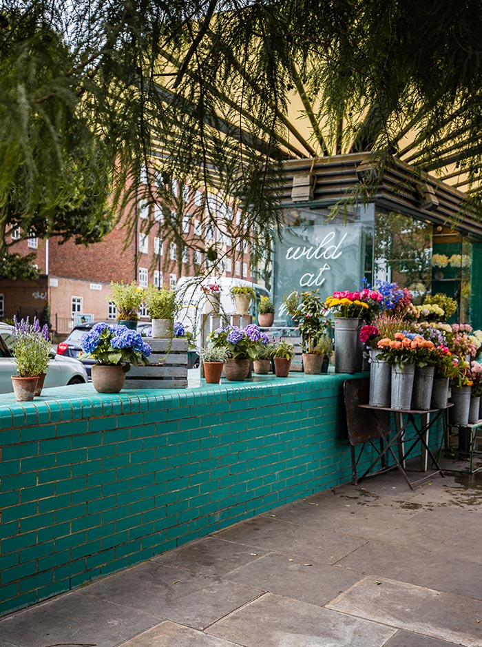 The exterior of Wild at Heart, a florist in Notting Hill, London, features a vibrant display of colorful flowers in various pots and metal buckets. The teal-tiled wall and the canopy overhead add a charming touch to the quaint storefront, creating an inviting atmosphere for passersby.