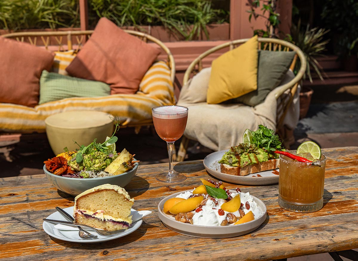 An inviting brunch setup at Wildes in Margate, featuring a variety of colorful and delicious dishes. The table is set with a bowl of roasted vegetables and greens, avocado toast, a slice of cake, and a dessert topped with peaches and whipped cream. Two refreshing cocktails accompany the meal. The background includes cozy seating with cushions and a lush, green ambiance, creating a perfect outdoor dining experience.