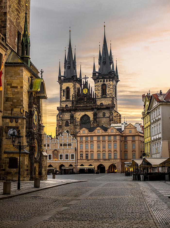 This image captures the iconic Church of Our Lady before Týn in Prague's Old Town Square, framed by the golden light of sunrise or sunset. The Gothic spires of the church tower dramatically over the surrounding historic buildings, including the Old Town Hall on the left, where Prague's famous Astronomical Clock can be seen. The cobblestone square in the foreground is empty, adding to the serene and timeless atmosphere of this historic and architectural landmark.