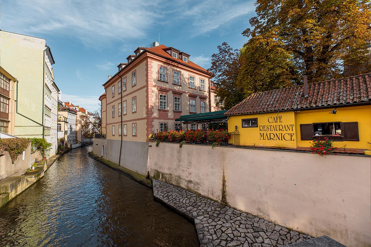 A picturesque canal view in Prague, featuring charming pastel-colored buildings lining the water, with a quaint yellow café adorned with vibrant flowers. The canal, reflecting the clear blue sky, adds a tranquil ambiance to this scene of "Little Venice" in the city.