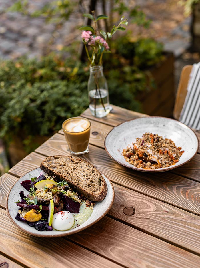 This image shows a beautifully set outdoor brunch table at Etapa, a popular café in Prague. The table features a rustic wooden surface with two plates of food: one with a slice of rustic bread, poached eggs, roasted vegetables, and grains, and another with a bowl of granola topped with fruit and yogurt. A glass of frothy coffee, likely a latte or cappuccino, sits next to a small vase with pink flowers, adding a touch of elegance to the cozy, natural setting.