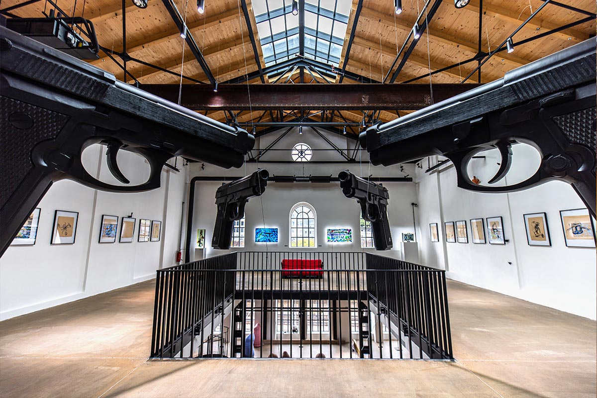 This image features an interior view of the "Musoleum" art installation by David Černý, located in Prague. The space is dominated by large, suspended sculptures of black pistols pointing towards each other, creating a striking and intense atmosphere. The gallery's bright white walls and natural light streaming through the large windows contrast with the dark, imposing figures of the guns. Framed artworks hang on the walls, adding an additional layer of visual interest to the modern, industrial setting.