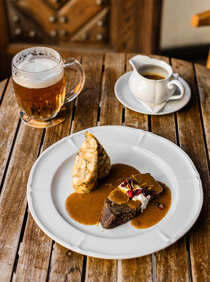 This image showcases a traditional Czech dish called "Svíčková" served on a rustic wooden table. The plate features tender beef topped with a creamy, rich sauce and garnished with cranberries and orange zest, accompanied by a slice of bread dumpling (knedlík). Beside the dish, there's a frothy glass of Czech beer and a small white pitcher of extra sauce, enhancing the meal's presentation. The warm, cozy setting highlights the hearty and comforting nature of this classic Czech cuisine.