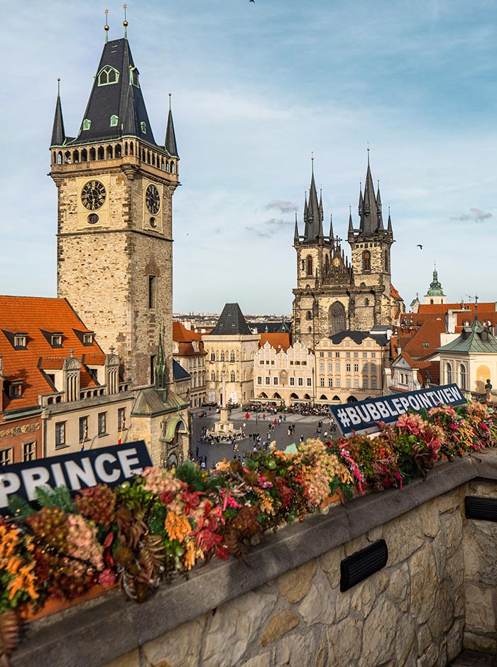 This image presents a breathtaking view from the terrace of U Prince, a popular spot in Prague. The vantage point overlooks the Old Town Square, with the Gothic spires of the Church of Our Lady before Týn dominating the background. The Old Town Hall tower and its famous Astronomical Clock are prominently featured on the left. The terrace is adorned with a vibrant floral arrangement, with signs reading "#BUBBLEPOINTVIEW" and "PRINCE" adding a modern touch to the historic setting. The scene captures the lively atmosphere and architectural beauty of one of Prague's most iconic locations.