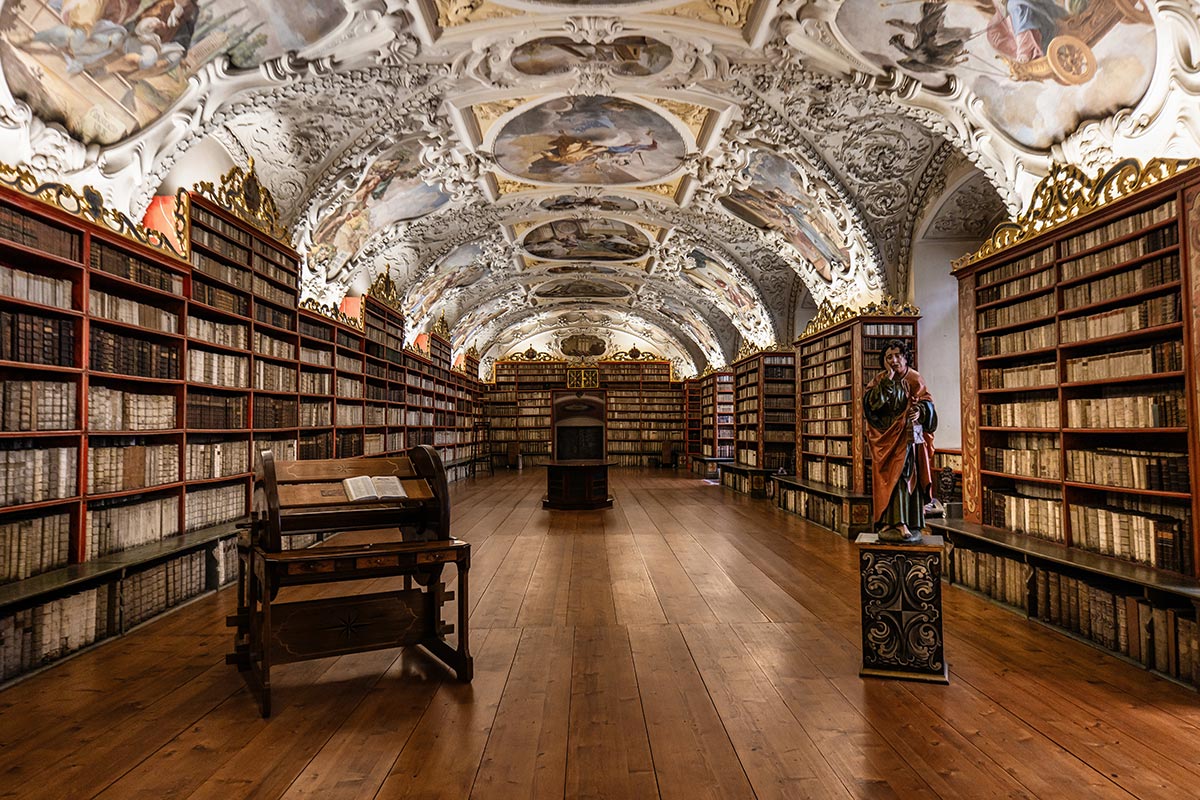 This image captures the Theological Hall at the Strahov Monastery in Prague, a stunning example of Baroque architecture. The hall is lined with floor-to-ceiling wooden bookshelves filled with ancient volumes, and the ornate ceiling is adorned with intricate frescoes depicting various religious and allegorical scenes. In the center of the room stands a large, antique reading desk with an open book, and a statue of a saint or religious figure is displayed on a pedestal to the right. The polished wooden floor adds warmth to the grand, scholarly atmosphere of the space.