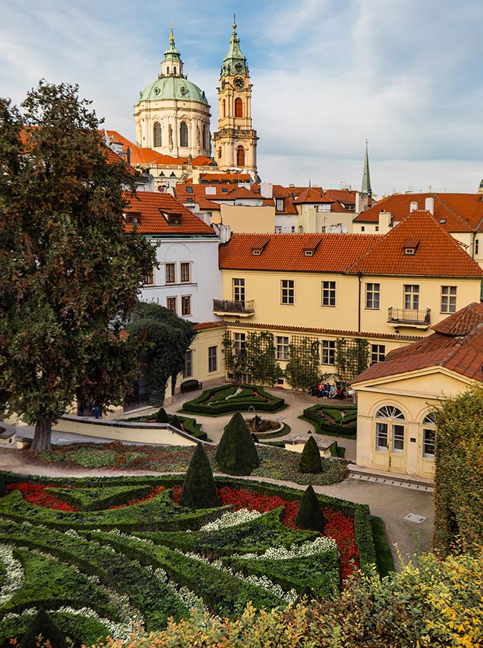 This image showcases the Vrtba Garden in Prague, a beautifully manicured Baroque garden located in the city's historic district. The garden features intricate, symmetrical patterns of hedges, vibrant flower beds, and carefully shaped topiary, all arranged on terraced levels. In the background, the twin towers of St. Nicholas Church rise above the red-tiled rooftops, adding to the scenic and architectural charm of the setting. The soft afternoon light enhances the rich colors and detailed landscaping of this tranquil urban oasis.