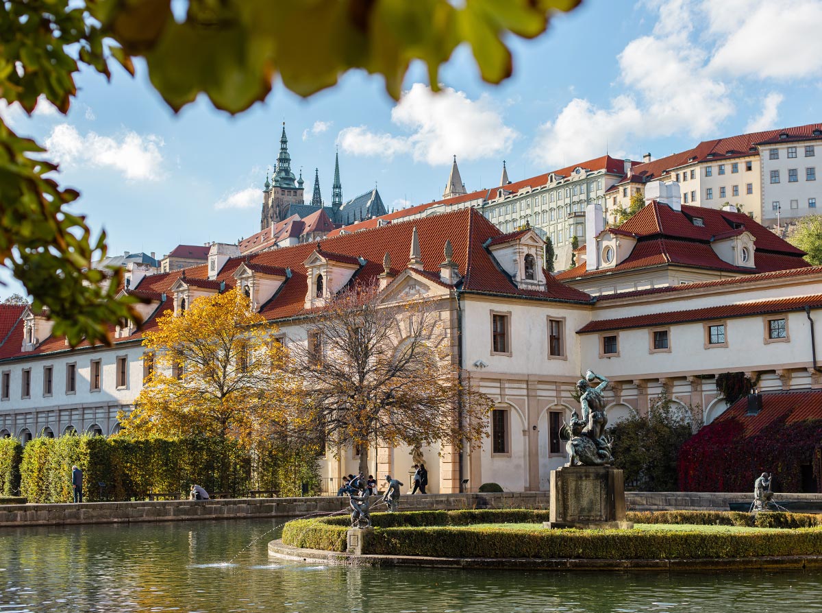 The image depicts Waldstein Garden in Prague, showcasing a serene pond with a small fountain and a statue in the center, surrounded by well-manicured hedges. In the background, there are historic buildings with red-tiled roofs, and the spires of St. Vitus Cathedral are visible in the distance, framed by autumnal trees and leaves. The scene captures the peaceful elegance of the garden and the rich architectural heritage of Prague.