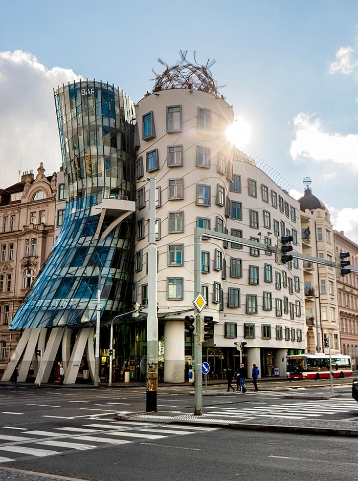 This image shows the Dancing House in Prague, Czech Republic, an iconic building with a unique, deconstructivist architectural design. The structure features two contrasting towers—one with a glass facade curving inward, resembling a woman in a dress, and the other with a more traditional, rectangular shape. The sun is shining behind the building, casting a bright glow on its exterior. The street in the foreground includes a crosswalk and traffic signals, with a few pedestrians and a bus visible.