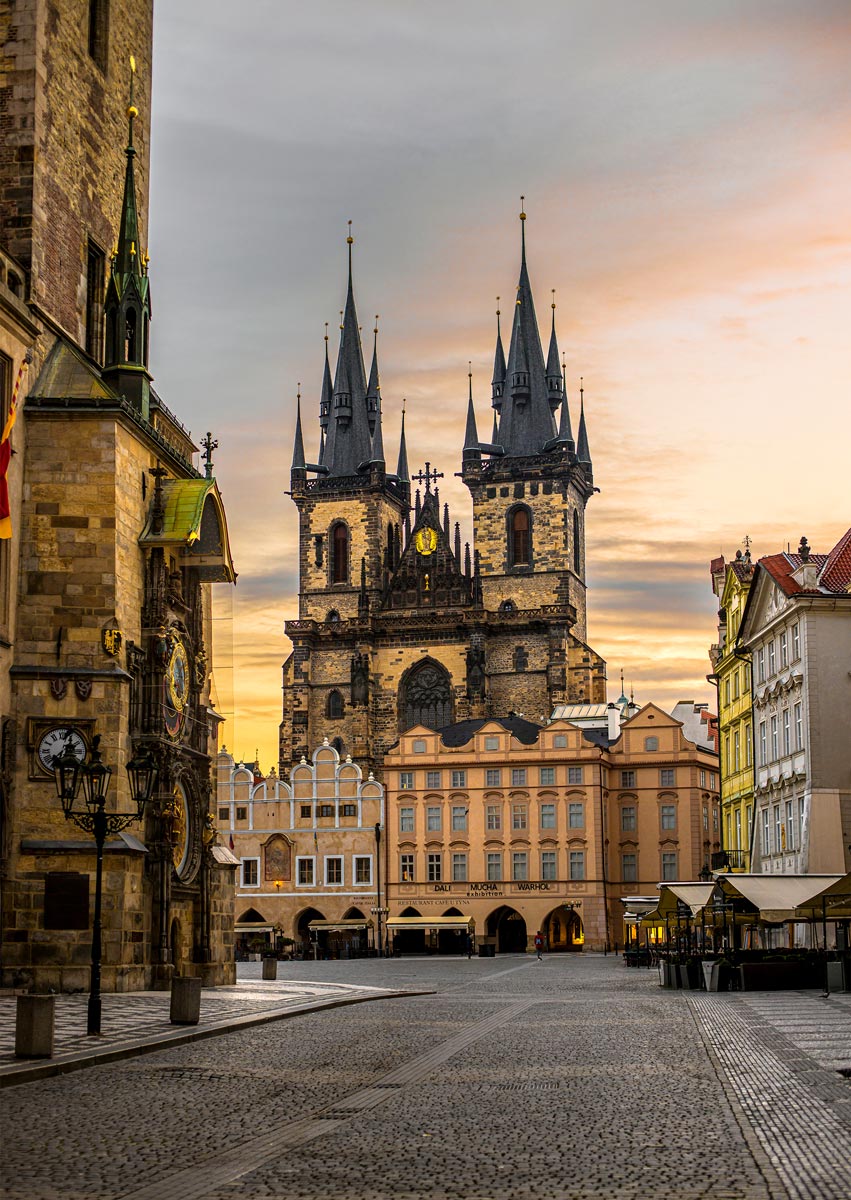 This image captures the iconic Church of Our Lady before Týn in Prague's Old Town Square, framed by the golden light of sunrise or sunset. The Gothic spires of the church tower dramatically over the surrounding historic buildings, including the Old Town Hall on the left, where Prague's famous Astronomical Clock can be seen. The cobblestone square in the foreground is empty, adding to the serene and timeless atmosphere of this historic and architectural landmark.