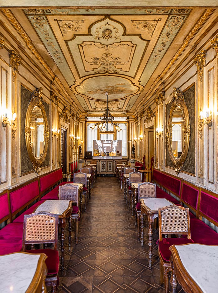 This image shows the elegant interior of Caffè Florian in Venice, Italy, one of the oldest coffee houses in the world. The narrow room is adorned with ornate, gilded mirrors, intricate wall moldings, and a richly decorated ceiling. Red velvet seating lines both sides, paired with marble-topped tables and classic wooden chairs, creating an atmosphere of historical grandeur and luxury.