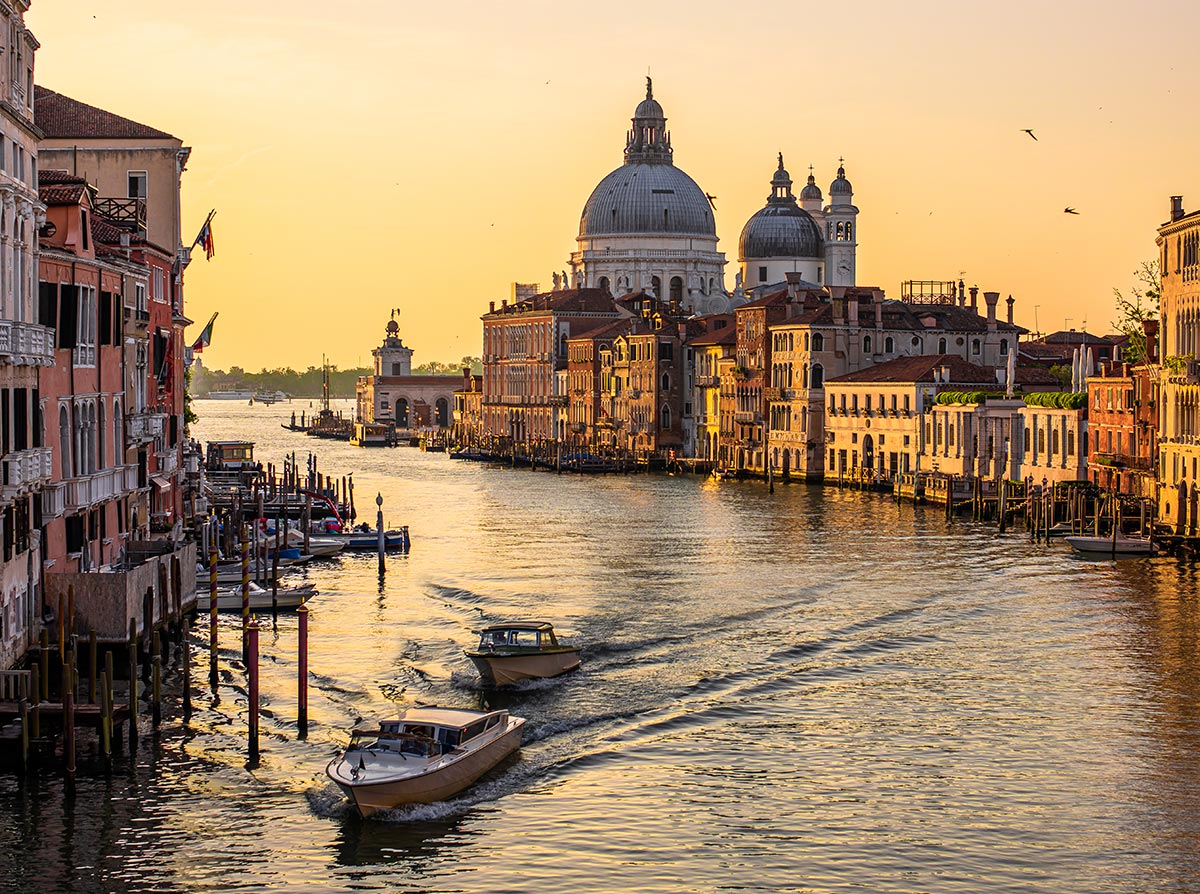 This image captures the iconic Grand Canal in Venice, Italy, during sunset. The scene features boats gliding through the canal, flanked by historic buildings bathed in golden light. In the background, the grand dome of the Basilica di Santa Maria della Salute dominates the skyline, creating a picturesque view of Venice’s waterways and architecture. The calm water reflects the warm hues of the setting sun, enhancing the city's timeless beauty.