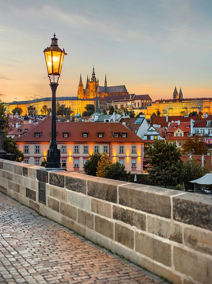 This image captures a stunning evening view from Charles Bridge in Prague, looking towards Prague Castle. The scene is illuminated by the soft glow of a traditional street lamp in the foreground, while the historic rooftops and buildings below are bathed in warm, golden light as the sun sets. In the background, the majestic St. Vitus Cathedral towers over the city, adding to the enchanting atmosphere of this iconic and picturesque location. The cobblestone pathway on the bridge adds texture and a sense of timelessness to the composition.