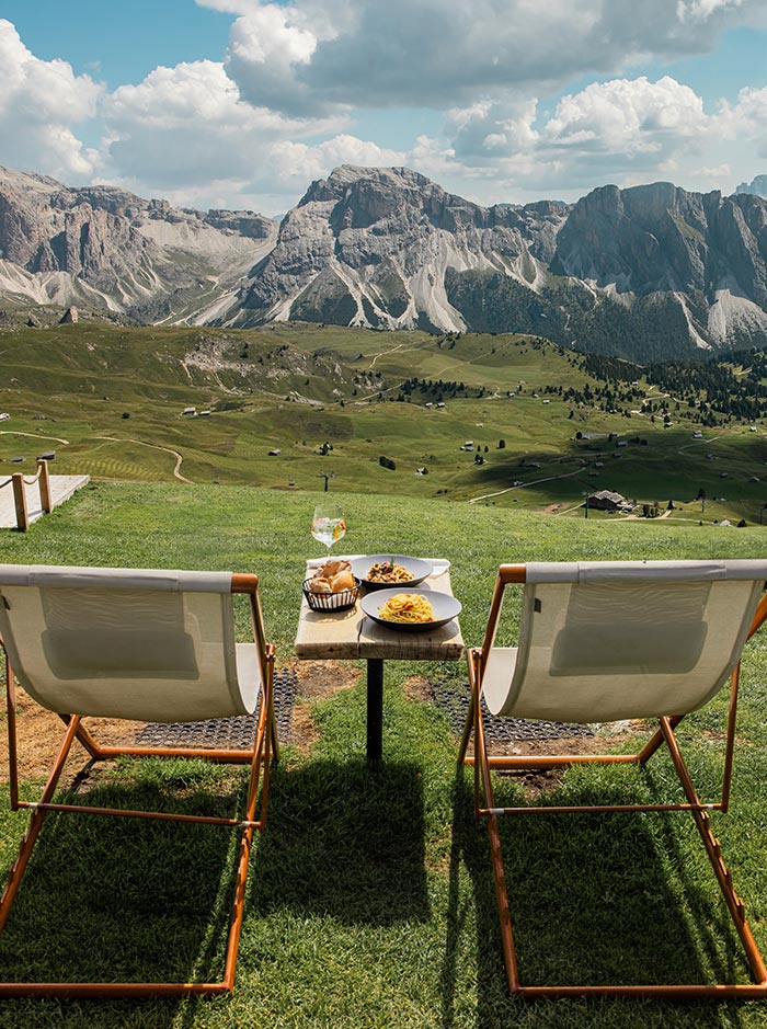 This image shows two lounge chairs facing a table set with plates of food and a glass of drink, all positioned on a grassy slope. The backdrop features a breathtaking view of the rugged Dolomite mountains and rolling green hills under a partly cloudy sky. The scene offers a perfect blend of relaxation, fine dining, and panoramic mountain views, ideal for a peaceful outdoor meal in nature.