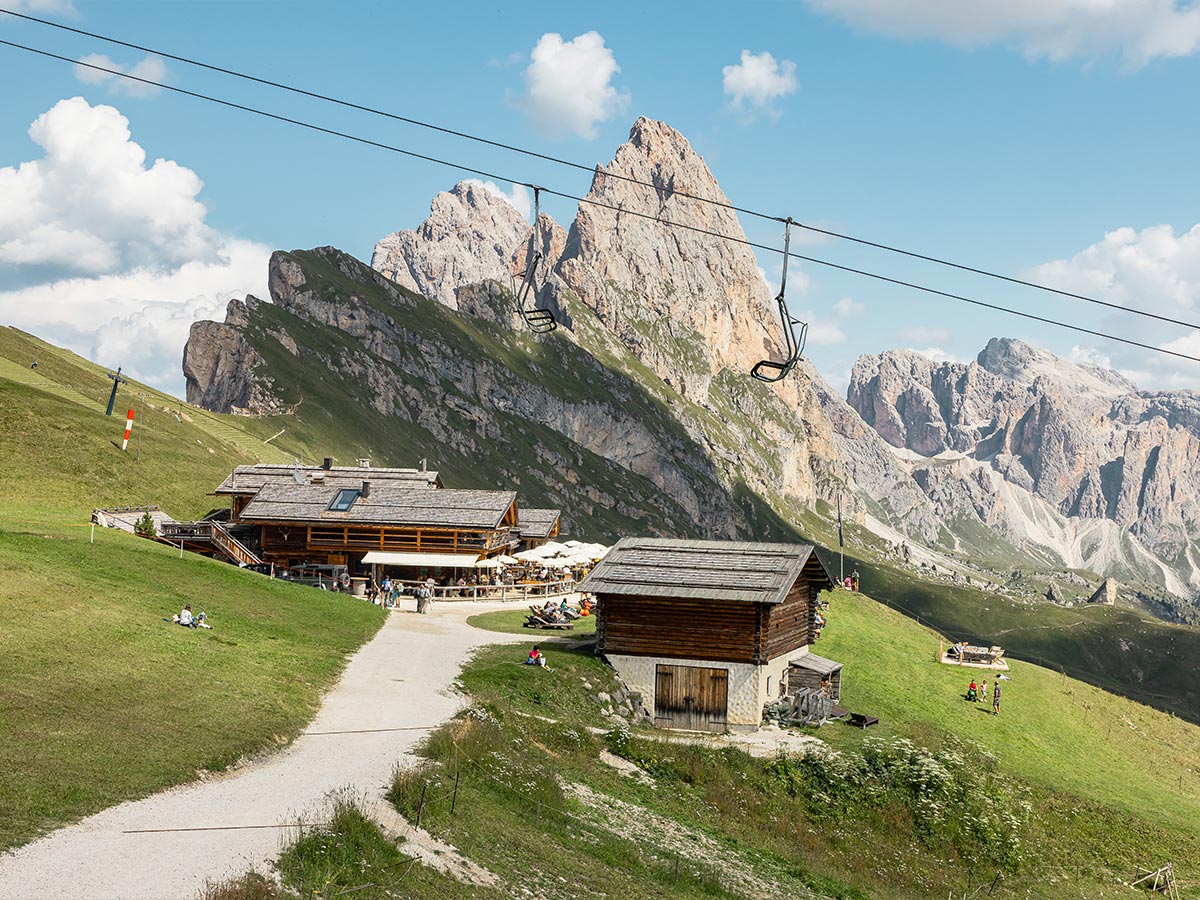 This image features a cozy mountain hut, "Baita Sofie Hütte," set in a grassy alpine landscape with the rugged Seceda peaks towering in the background. Visitors are seen relaxing outside, and a ski lift chair hangs above, suggesting this is a popular spot for both summer and winter activities. The wooden cabins blend harmoniously with the natural surroundings, offering a picturesque setting for rest and refreshments amid breathtaking mountain views.