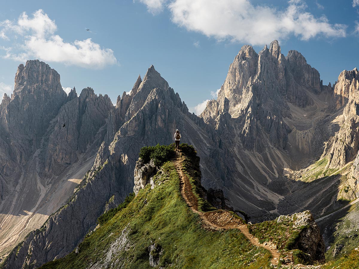 This image captures a hiker standing on a narrow mountain ridge, surrounded by the dramatic, jagged peaks of the Cadini di Misurina in the Dolomites. The sharp cliffs and rocky formations rise steeply on all sides, contrasting with the lush greenery along the hiking trail. The scene evokes a sense of adventure and awe, with the vast, rugged landscape under a bright, partly cloudy sky.