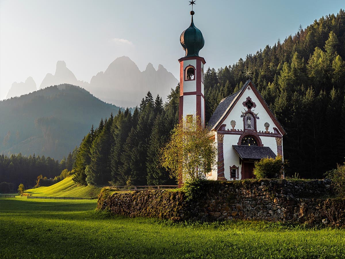 This image features the picturesque Church of St. John (San Giovanni) in Val di Funes, nestled against a backdrop of lush green fields, dense forests, and the towering, misty peaks of the Dolomites in the distance. The small, white chapel with its distinctive green onion dome is bathed in soft, early morning or late afternoon light, creating a peaceful and serene atmosphere. The surrounding landscape, with its rolling hills and vibrant greenery, adds to the idyllic charm of this iconic alpine location.