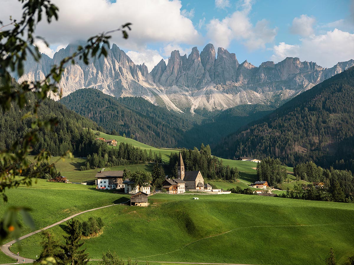 This image features the idyllic village of St. Magdalena in Val di Funes, with its iconic church nestled among lush green hills, set against the dramatic backdrop of the jagged peaks of the Dolomites. The scene captures the peaceful charm of this alpine landscape, with scattered farmhouses, winding roads, and dense forests. The towering mountains in the background provide a stunning contrast to the vibrant greenery of the valley, making this a quintessential Dolomites view.