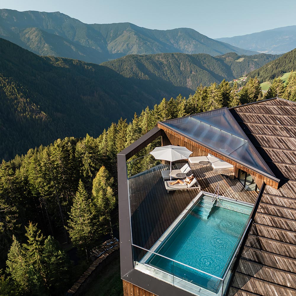 This image shows a stunning view of an elevated infinity pool and a deck with lounge chairs under an umbrella, overlooking a vast forested valley with mountain ranges in the distance. The scene captures a peaceful, luxurious retreat nestled in nature, offering both relaxation and breathtaking panoramic views of the surrounding Dolomites.






