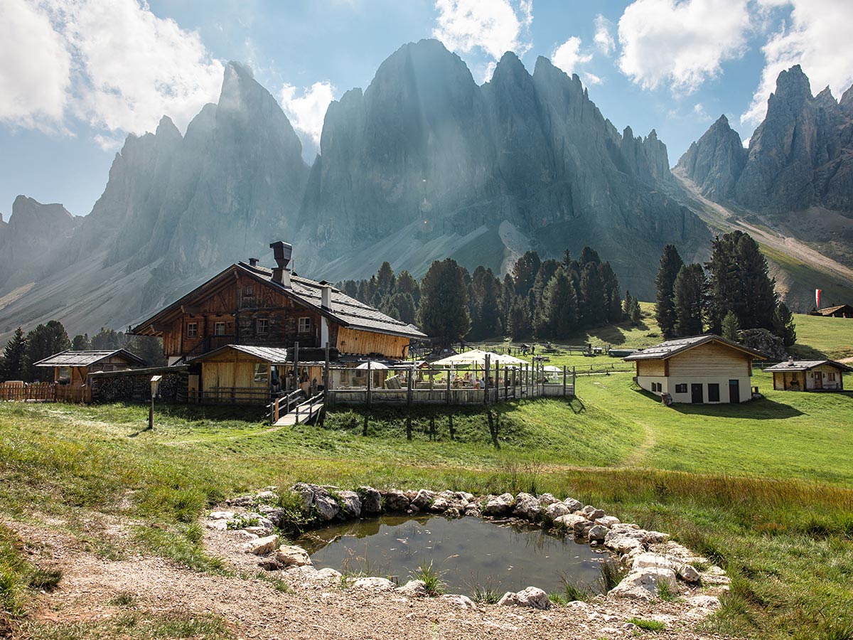 This image shows a charming alpine hut, Geisler Alm, set against the dramatic backdrop of the towering, jagged peaks of the Dolomites. The wooden structure is surrounded by green meadows, a small pond, and a few other buildings, creating a peaceful, rustic scene. Sunlight filters through the clouds, casting rays onto the rocky cliffs and lush landscape. The overall atmosphere is tranquil, offering a perfect retreat in the heart of nature.