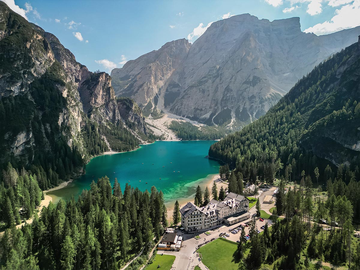 This image captures an aerial view of Lago di Braies (Pragser Wildsee) surrounded by lush forests and towering mountains in the Dolomites. The crystal-clear, turquoise waters of the lake contrast beautifully with the steep cliffs and dense greenery. A grand hotel sits at the edge of the lake, nestled among the trees, offering a tranquil and scenic retreat. The expansive landscape and serene atmosphere make this a perfect alpine getaway.