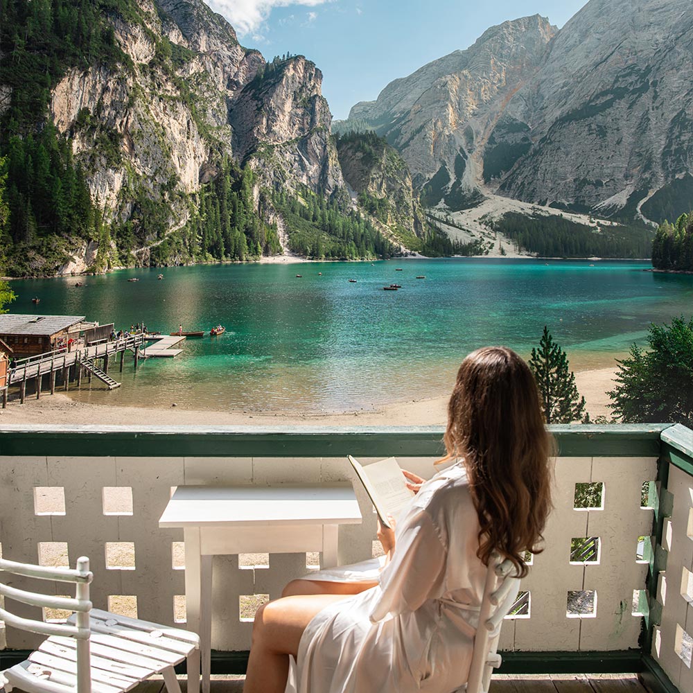 This image depicts a serene lakeside scene, with a woman in a white robe sitting on a balcony while reading a book. She overlooks the crystal-clear waters of a tranquil lake, surrounded by steep, forested mountains and rocky cliffs. Small boats can be seen on the water, and a rustic dock extends into the lake, enhancing the peaceful, idyllic atmosphere of this alpine setting.






