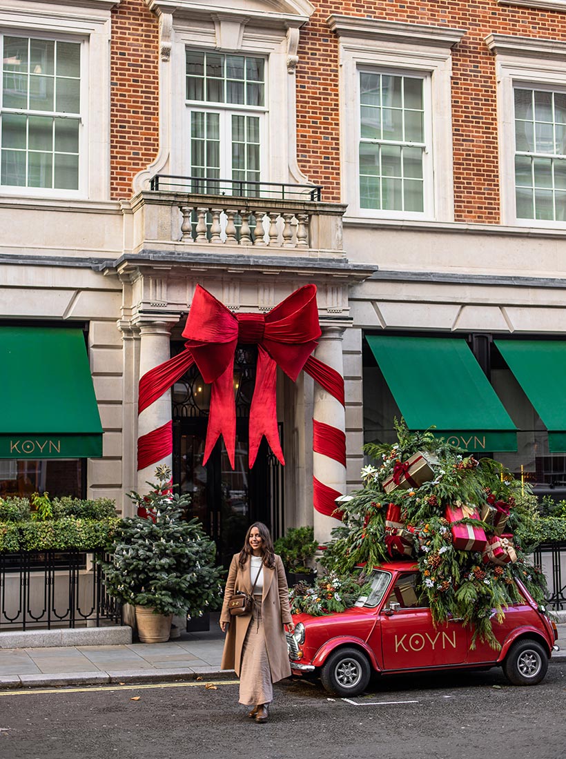 This image shows the festive exterior of KOYN restaurant, adorned with large holiday decorations. Two candy cane pillars wrapped with a giant red ribbon and bow frame the entrance. A red vintage car, covered with greenery and stacked with presents on the roof, is parked outside, adding to the seasonal charm. A woman in a beige coat and crossbody bag walks past the display, which is surrounded by small Christmas trees and greenery, creating a joyful holiday scene.