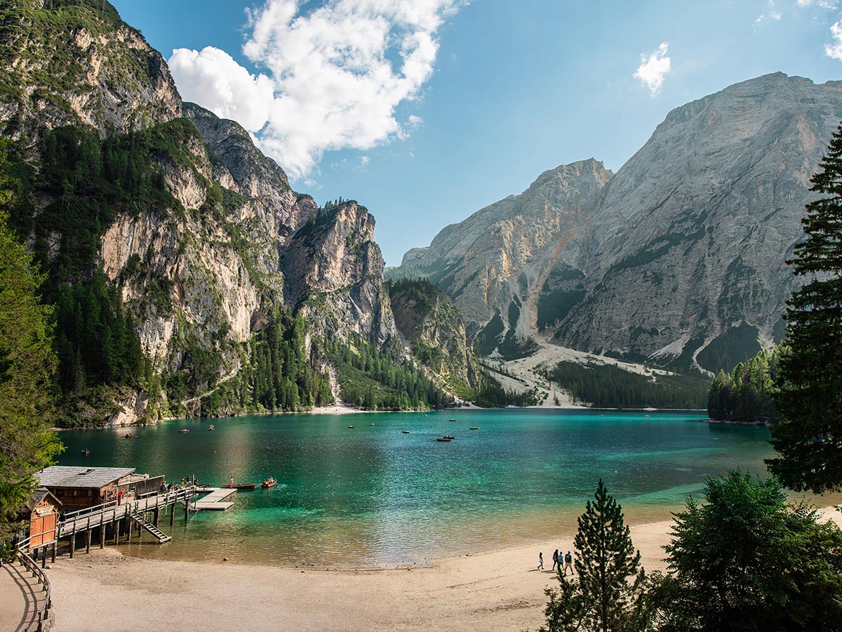 This image showcases the pristine Lago di Braies (Pragser Wildsee), with its clear turquoise waters surrounded by towering, forested mountains and steep rocky cliffs in the Dolomites. A wooden dock extends into the lake, where small boats float gently on the water. A sandy shore invites visitors to stroll along the lake, enjoying the peaceful alpine scenery under a bright sky. The combination of water, mountains, and greenery creates a picture-perfect natural landscape.